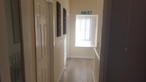 A hallway with a door on the left, a window with blinds, and a green exit sign above the window.