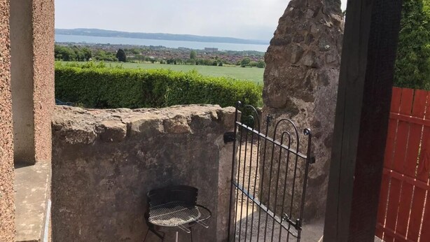 A stone wall with a metal gate and a chair, overlooking a green landscape and a distant body of water.