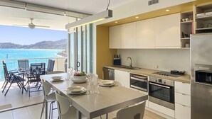 A modern kitchen with a view of the sea, a dining area with chairs and a table, and a built-in oven and sink. - 1 Bright Point Apartment 2305 (Nelly Bay)