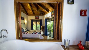 A bathroom with a large bathtub, wooden ceiling beams, a bed with a colorful quilt, and a view of a bedroom through a mirror. - Magnetic Cottage (Horseshoe Bay)