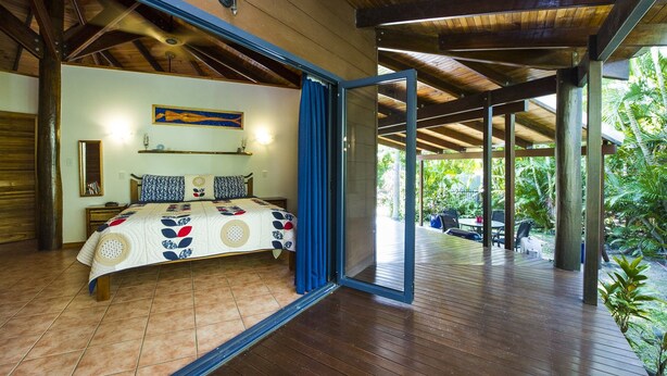 A bedroom with a bed, a nightstand, a wall-mounted shelf, and a large window with blue curtains. - Magnetic Cottage (Horseshoe Bay)