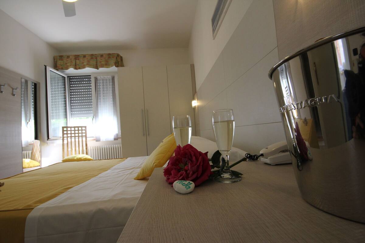A hotel room with a bed, a bedside table with a champagne glass, a flower, a telephone, and a minibar.