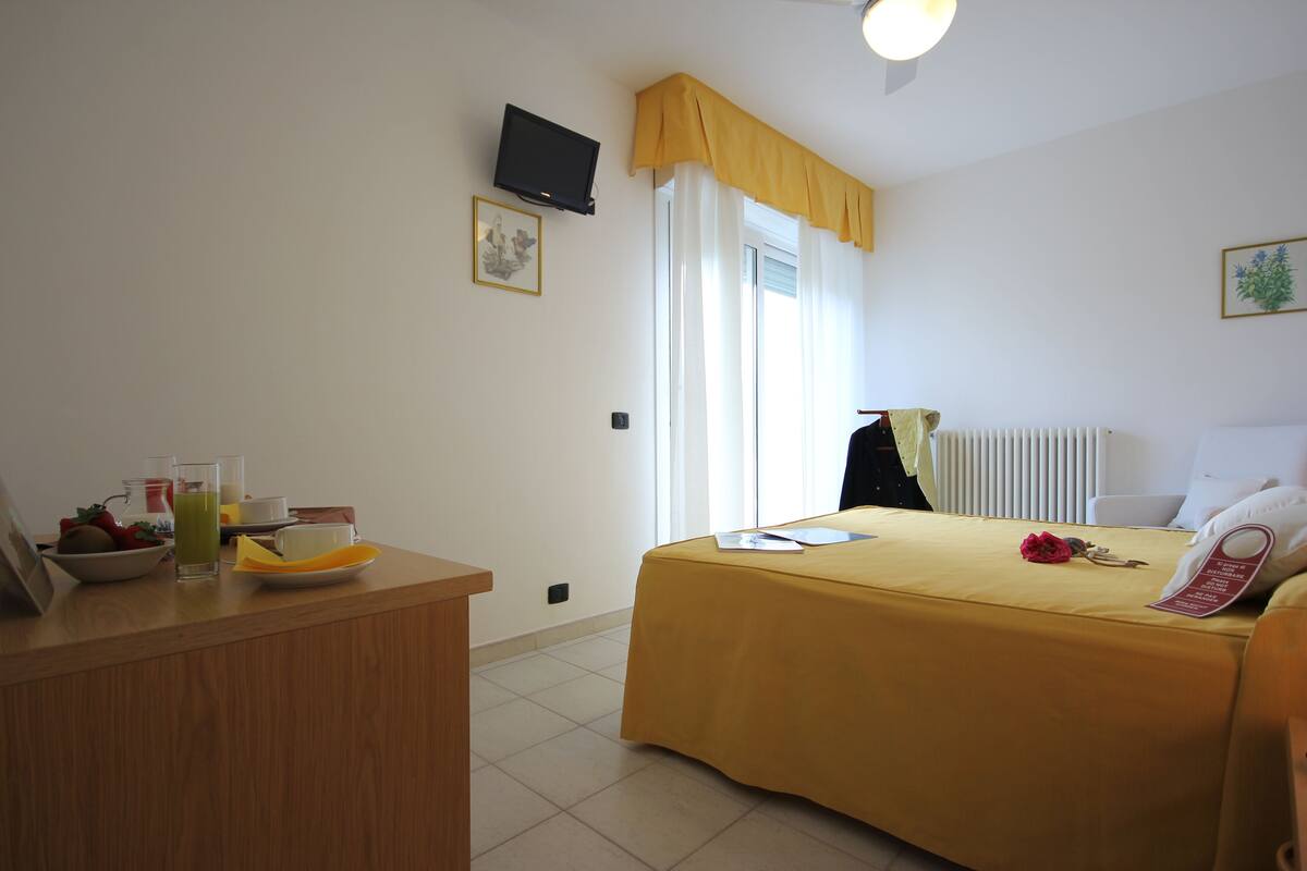 A hotel room with a bed, a desk with a telephone, a window with curtains, and a radiator.