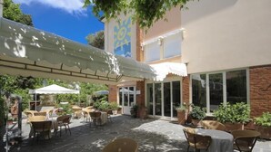 An outdoor patio area with tables and chairs, potted plants, and a building with a sun mural. - Hotel Miramare (Forte dei Marmi)
