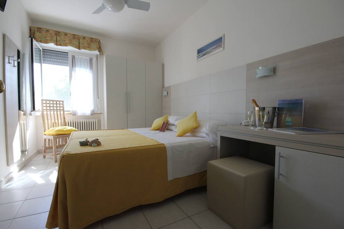 A hotel room with a bed, a desk, a chair, a radiator, and a window with curtains.