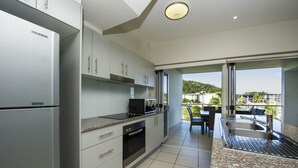 A modern kitchen with a built-in refrigerator, oven, and microwave. It features a large window overlooking a scenic view. - Beachside Apartment 14 (Nelly Bay)