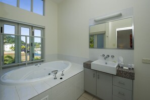 A modern bathroom with a large bathtub, a sink, and a mirror. There is a window providing a view of a pool and buildings. - Beachside Apartment 14 (Nelly Bay)