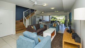A modern living room with a brown sofa, blue armchairs, and a white coffee table. A staircase leads to a kitchen area with a dining table and chairs. - Beachside Apartment 14 (Nelly Bay)