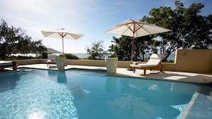 A clear swimming pool with lounge chairs and umbrellas, surrounded by trees and a view of the sea. - Lotus House (Horseshoe Bay)