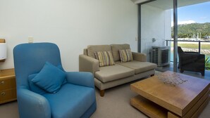 En stue med en blå lænestol, en beige sofa og et træbord. Der er en glasdør, der fører ud til en balkon med udsigt over vand og både.