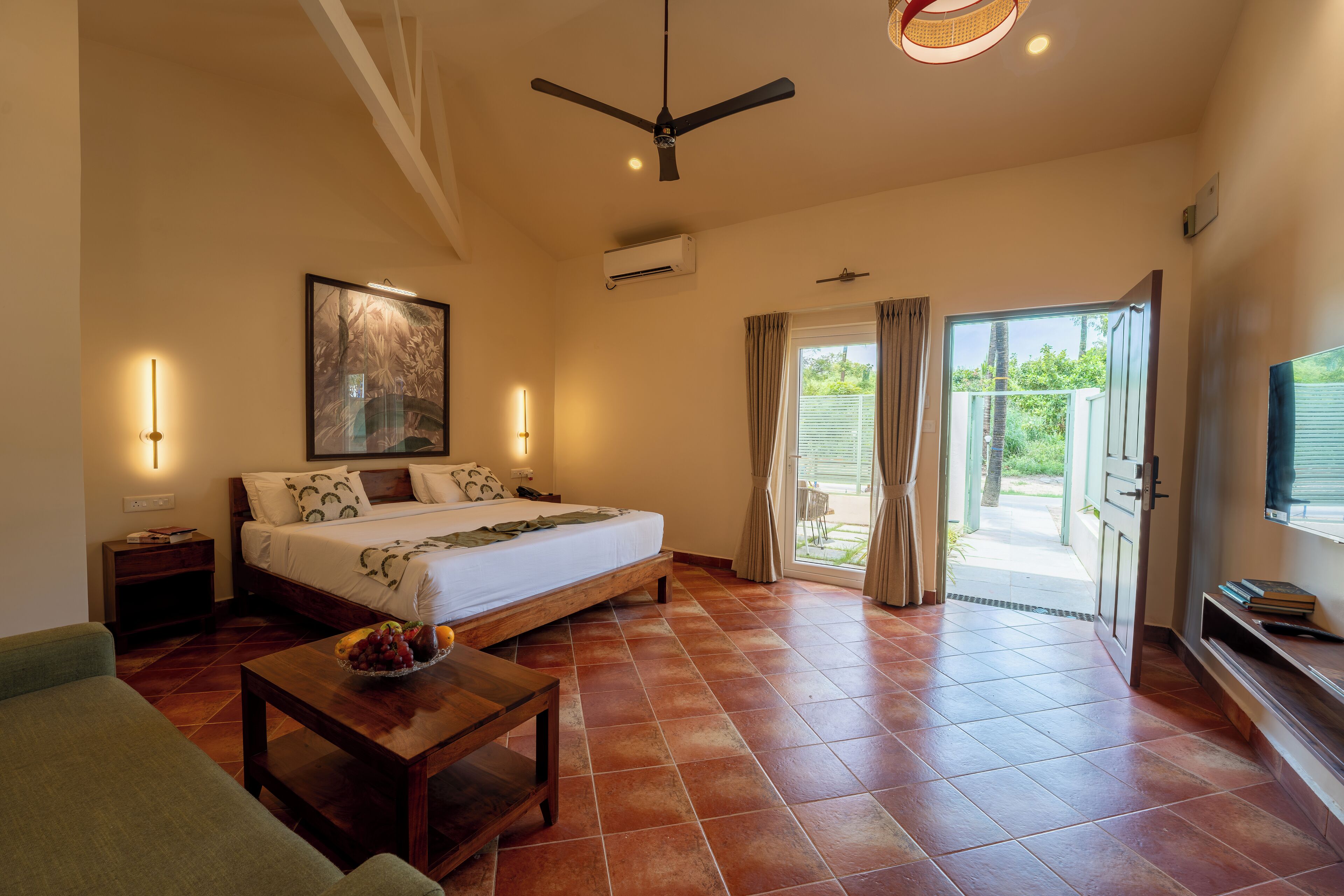 A spacious bedroom with a bed, a ceiling fan, a nightstand, a bedside table with a fruit bowl, and a view of the outside through a sliding glass door.