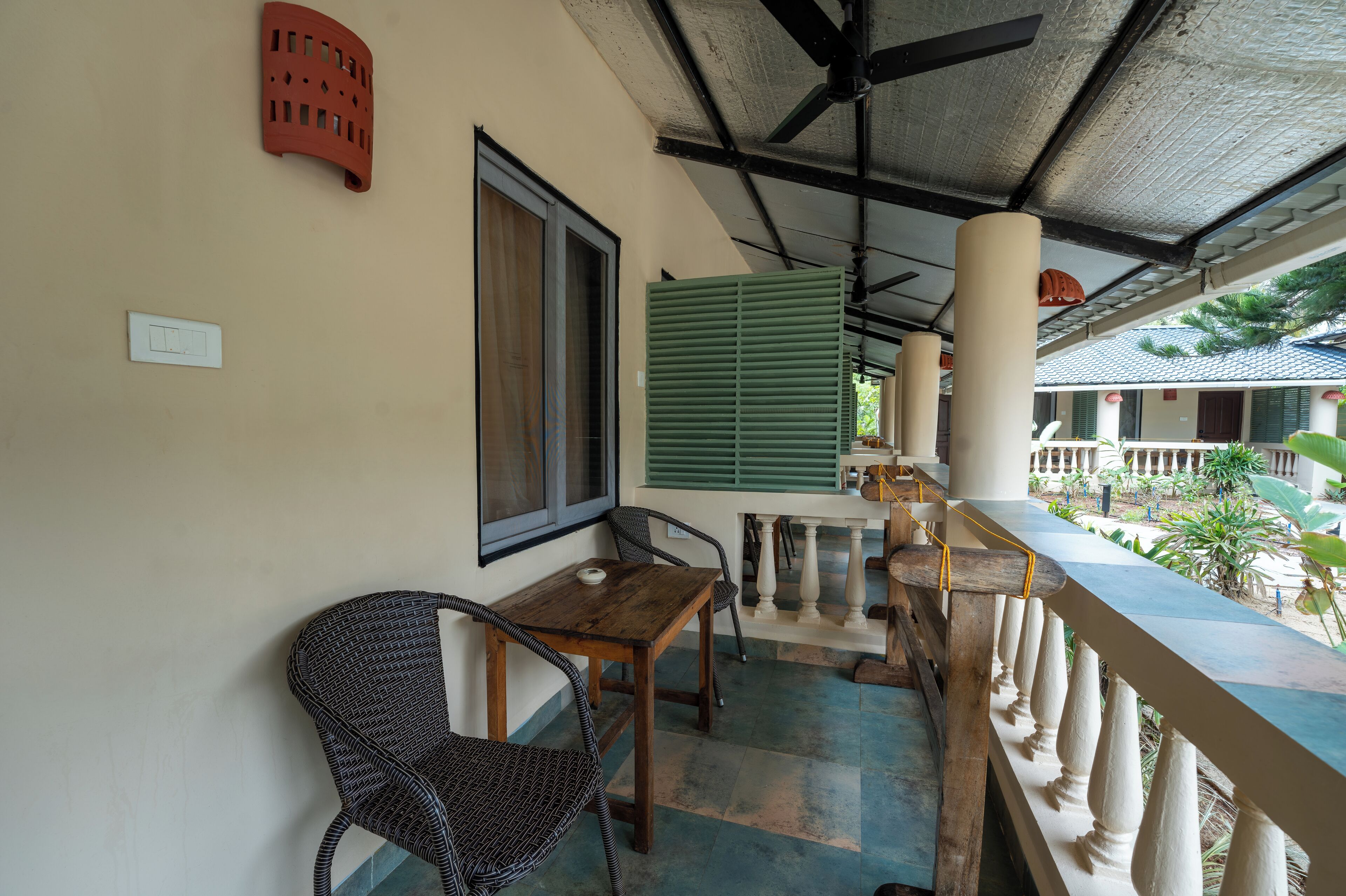 A balcony with a wooden table and chairs, a green shutter, and a view of a garden.