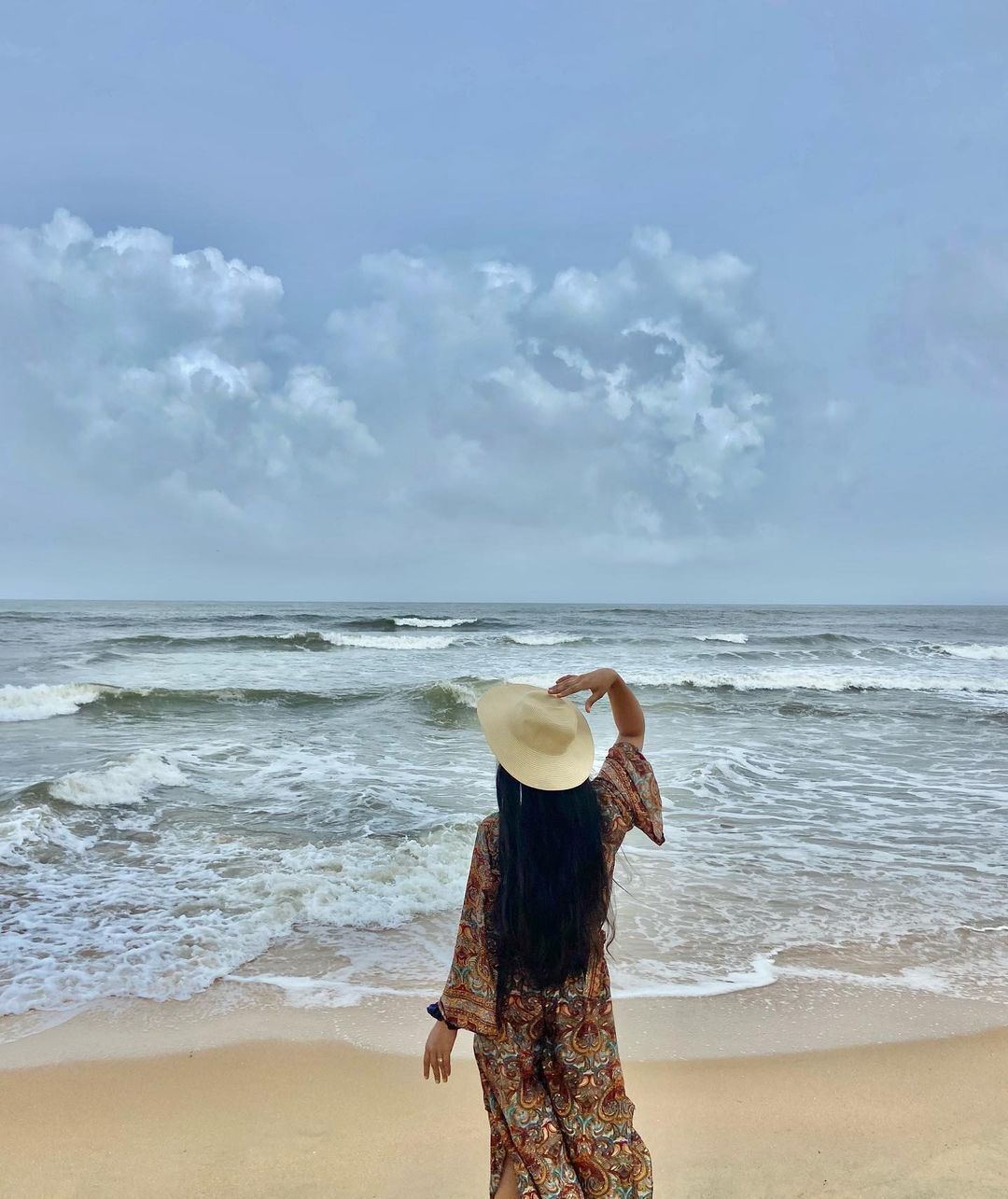 A person standing on a beach facing the ocean, wearing a patterned dress and a hat.