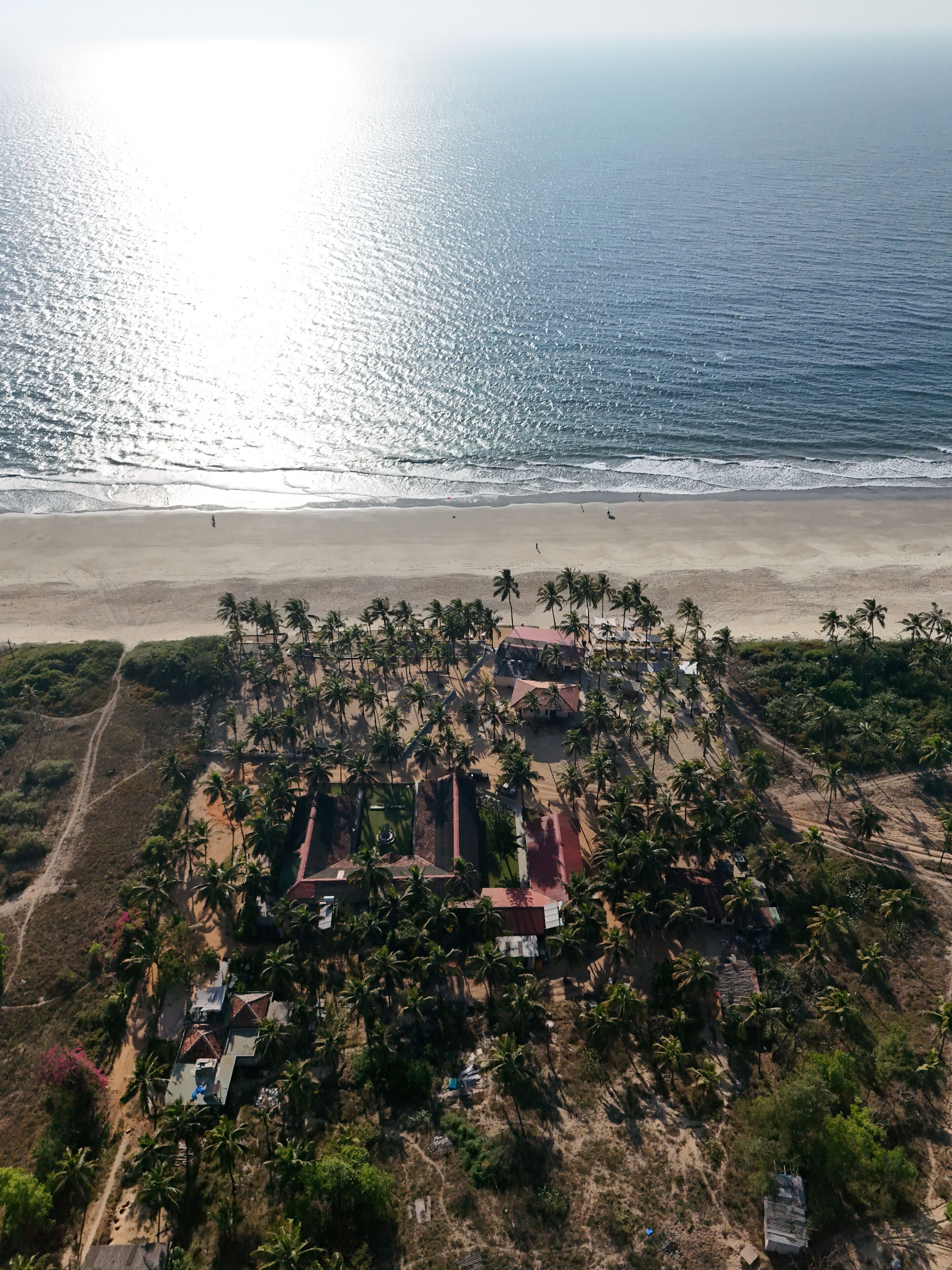 An aerial view of a coastal area with a beach, palm trees, and several buildings with red roofs.