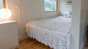 A bedroom with a large bed covered in a white lace blanket, a window with white curtains, and a wall-mounted light fixture. - Soltvedts Cabin - Bjønnes (Porsgrunn)