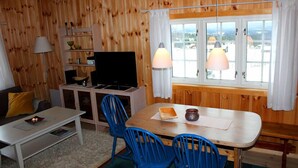 A cozy room with wooden walls, a television, a dining table with blue chairs, and a window with curtains.
