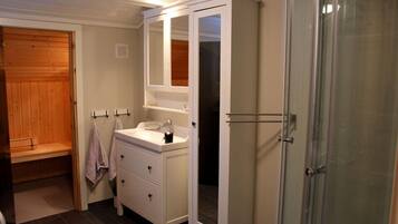 Um banheiro com porta de madeira, toucador branco com espelho e box de vidro.