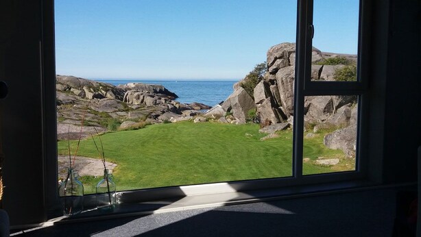 A room with a large window offering a view of a rocky coastline and grassy area. - Haver Cabin - Sirevåg (Ha)