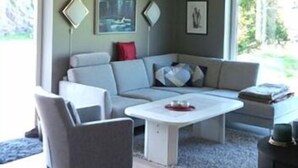 A modern living room with a grey sofa, a white coffee table, and a colorful rug. There are framed pictures on the wall and a window providing a view of the outdoors. - Haver Cabin - Sirevåg (Ha)