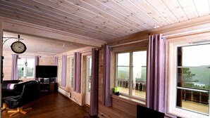Interior de una cabaña con paredes de madera, una mesa de comedor, un televisor y ventanas grandes que dan a un paisaje pintoresco.