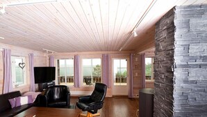 Una sala moderna con techo de madera, pared decorativa de piedra, televisor de pantalla plana, sillones de cuero, mesa de centro de madera y ventanas grandes con cortinas.