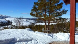 Eine Holzterrasse mit verschneiter Landschaft, einem großen Baum und einem Bergpanorama im Hintergrund.