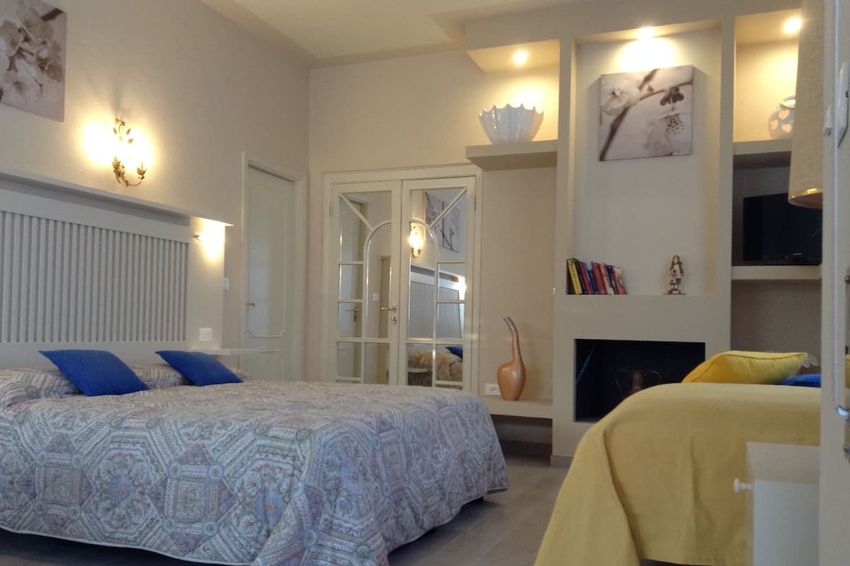 A bedroom with a large bed, a bedside table, a mirror, a vase, and a fireplace.