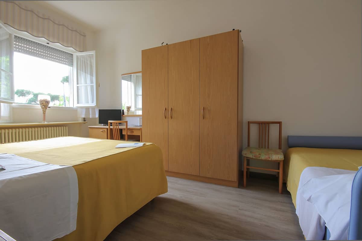 A bedroom with two beds, a wooden wardrobe, a desk with a chair, and a window with curtains.