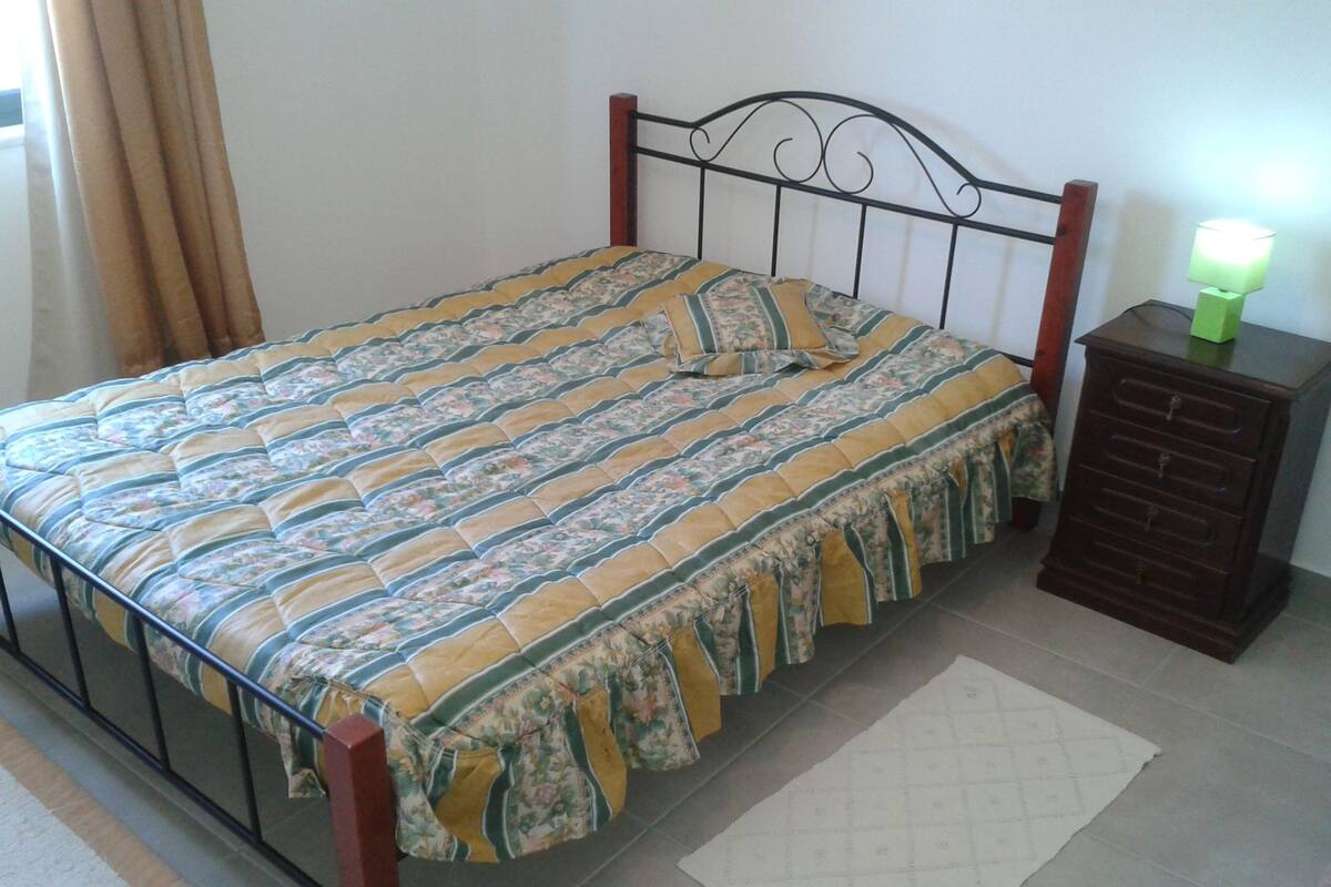 A single bed with a floral-patterned bedspread and a matching pillow, a wooden headboard, a small wooden nightstand with a green lamp, and a white rug on a tiled floor.