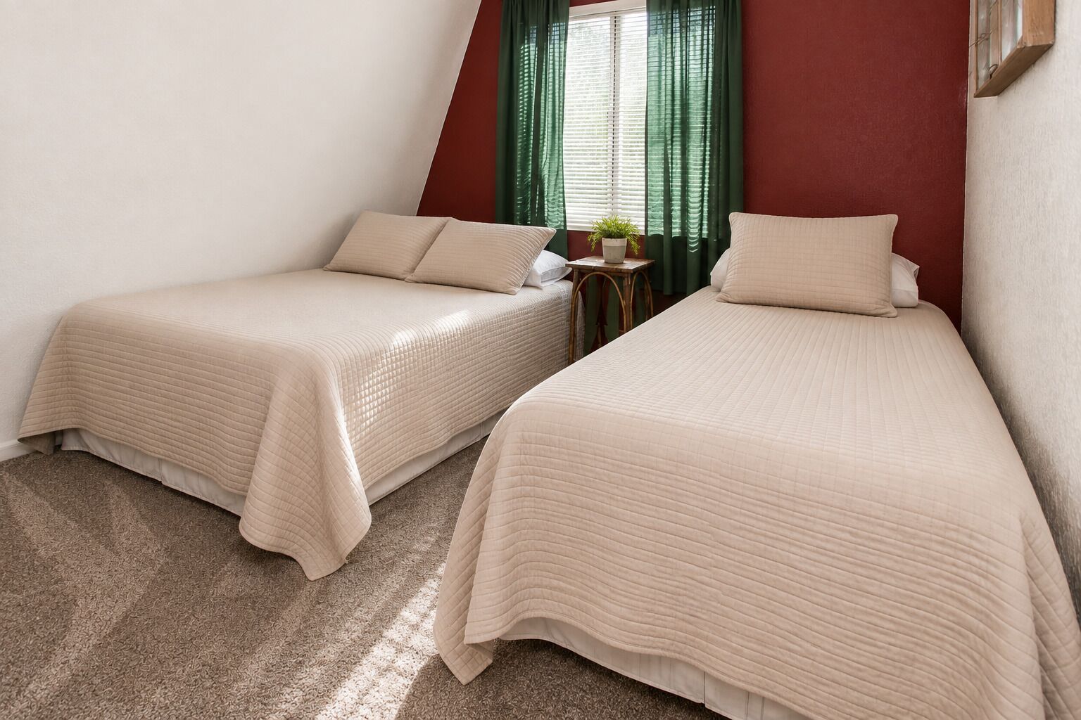 Dos camas individuales con ropa de cama color beige en una habitación con una pared de acento roja, una mesita con una planta y una ventana con cortinas verdes.