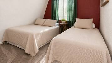Dos camas individuales con ropa de cama color beige en una habitación con una pared de acento roja, una mesita con una planta y una ventana con cortinas verdes.