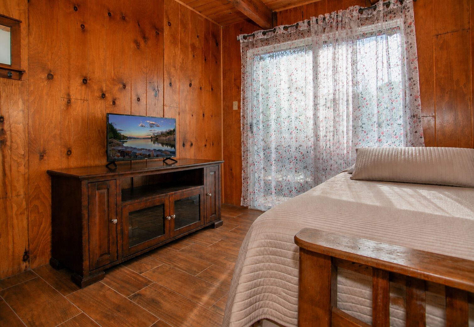 A wooden-paneled room with a bed, a TV on a cabinet, and a window with floral curtains.