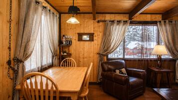 A cozy cabin interior with a wooden dining table, chairs, a leather sofa, and a lamp.