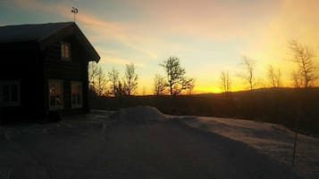 A log cabin with a sloped roof and a chimney, surrounded by snow-covered ground and leafless trees, during sunset.
