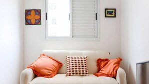 A room with a sofa, two pillows, a window with blinds, and two framed pictures on the wall. - Apartamento 2 dorms Metrô Saúde (São Paulo)