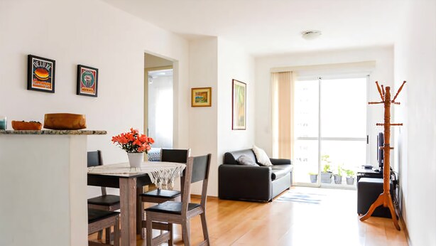 A dining area with a wooden table and chairs, a vase with flowers, and a sofa in the background. - Apartamento 2 dorms Metrô Saúde (São Paulo)