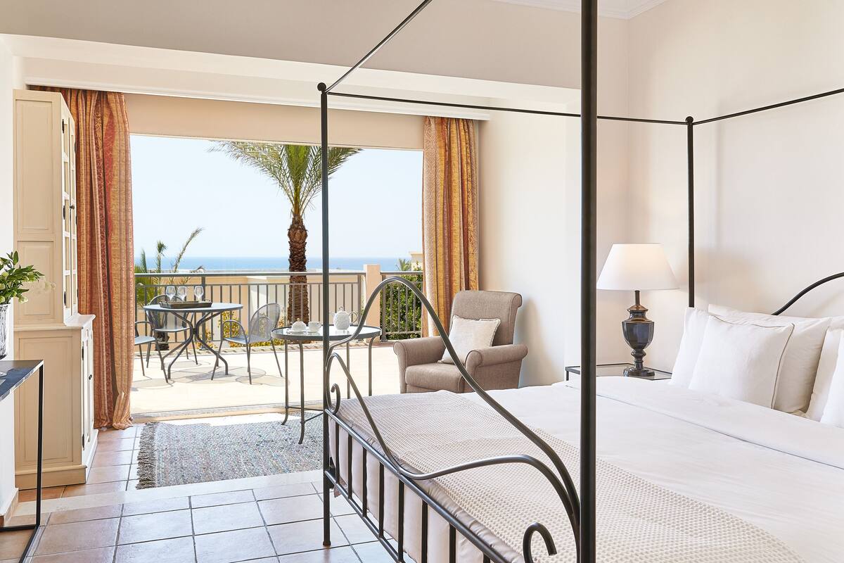 Una cama con dosel con vistas al mar, un balcón con mesa y sillas, y una lámpara en la mesita de noche.