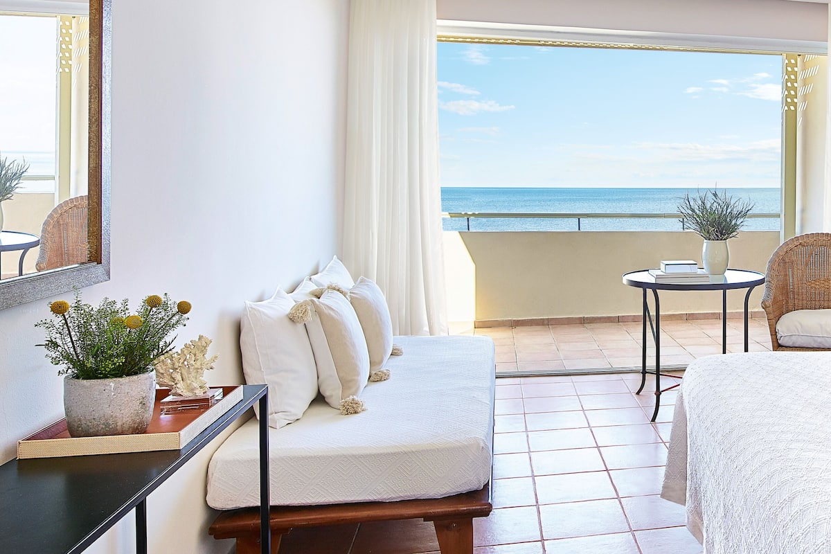Una habitación de hotel con una cama, una mesita con una planta, una silla y vista al mar.