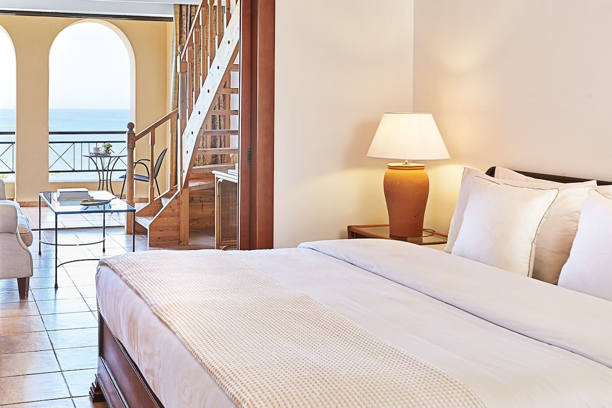 Habitación de hotel con cama, mesita de noche con lámpara, una silla y vista al mar desde el balcón.