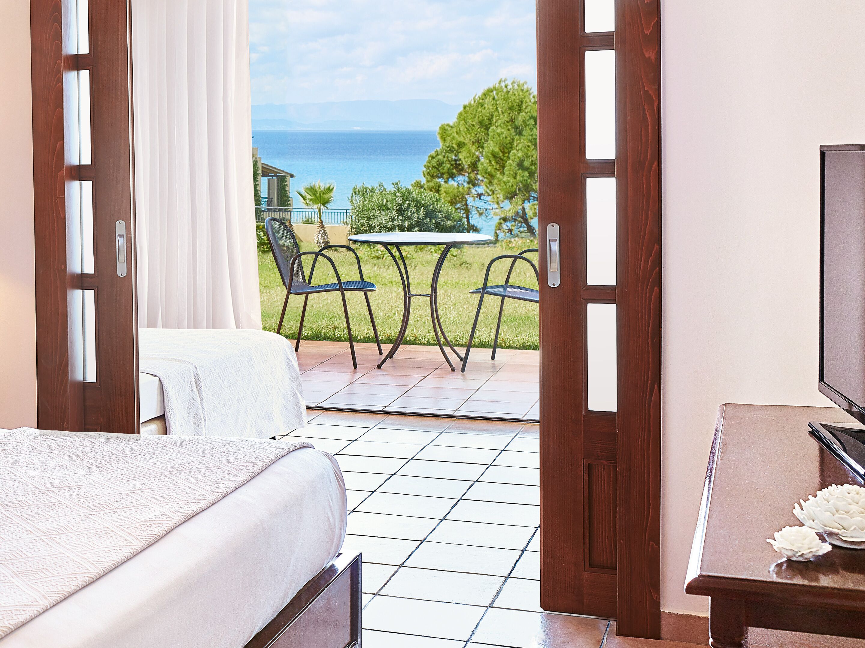 A hotel room with a bed, a TV, and a view of the sea through an open door.