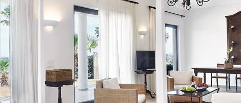 A spacious hotel room with a large bed, a television, wicker furniture, a dining table with fruit, and a view of the outdoors.