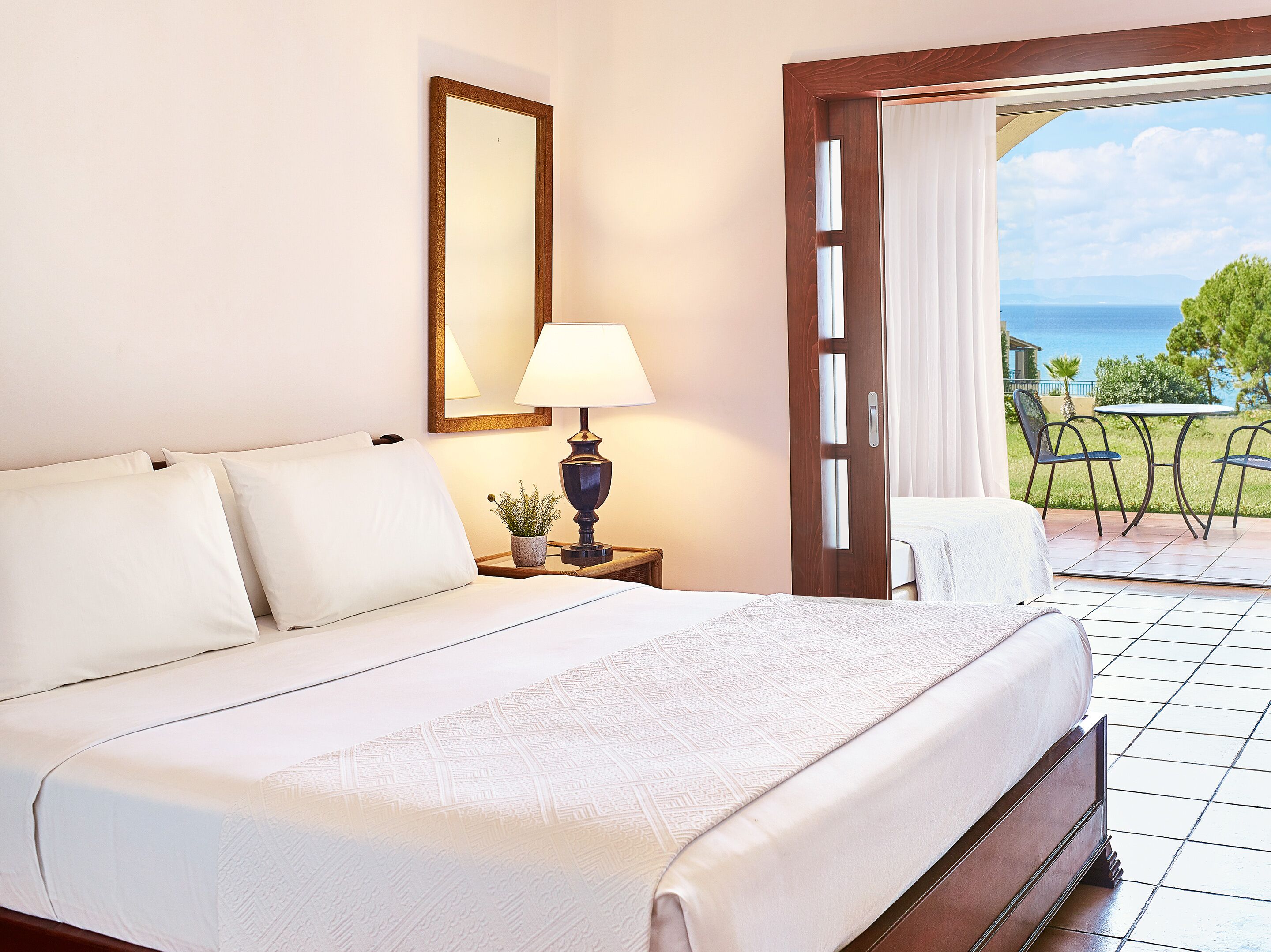 Une chambre d’hôtel avec un lit, une table de chevet avec une lampe, un miroir et une vue sur la mer par une porte ouverte.