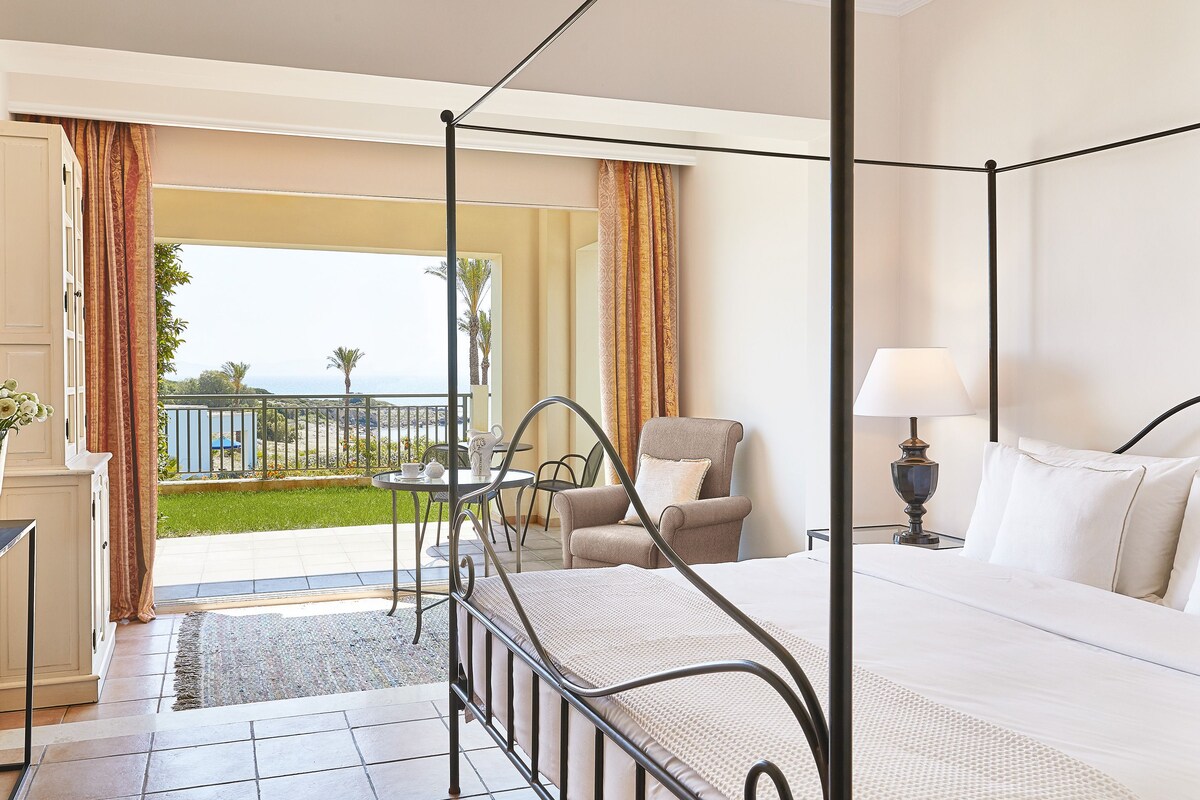 Una cama con dosel con vistas al mar, un balcón con mesa y sillas, y una lámpara en la mesita de noche.