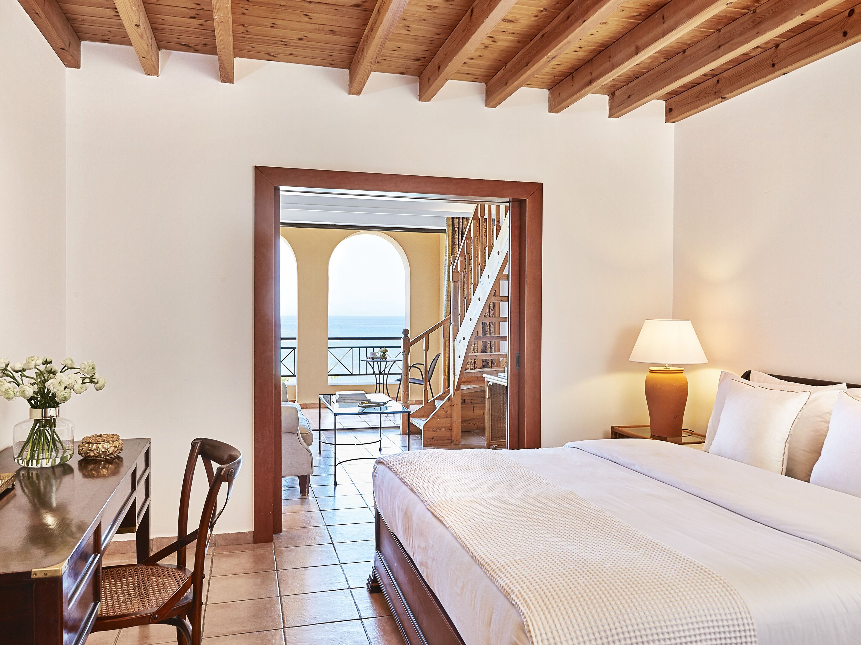 Un dormitorio con una cama grande, un escritorio con silla, una lámpara y vista al mar por un arco.