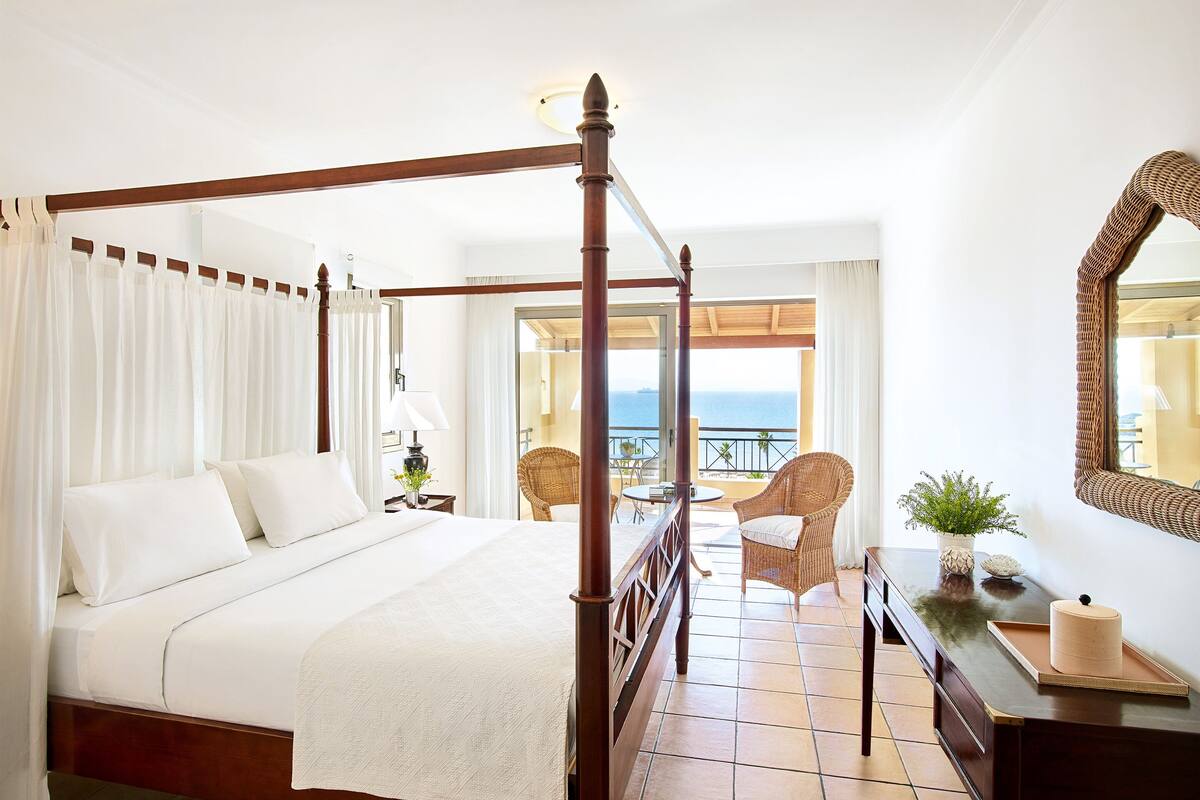 Una cama con dosel con sábanas blancas, un cabecero de madera y vistas al mar desde un balcón abierto.