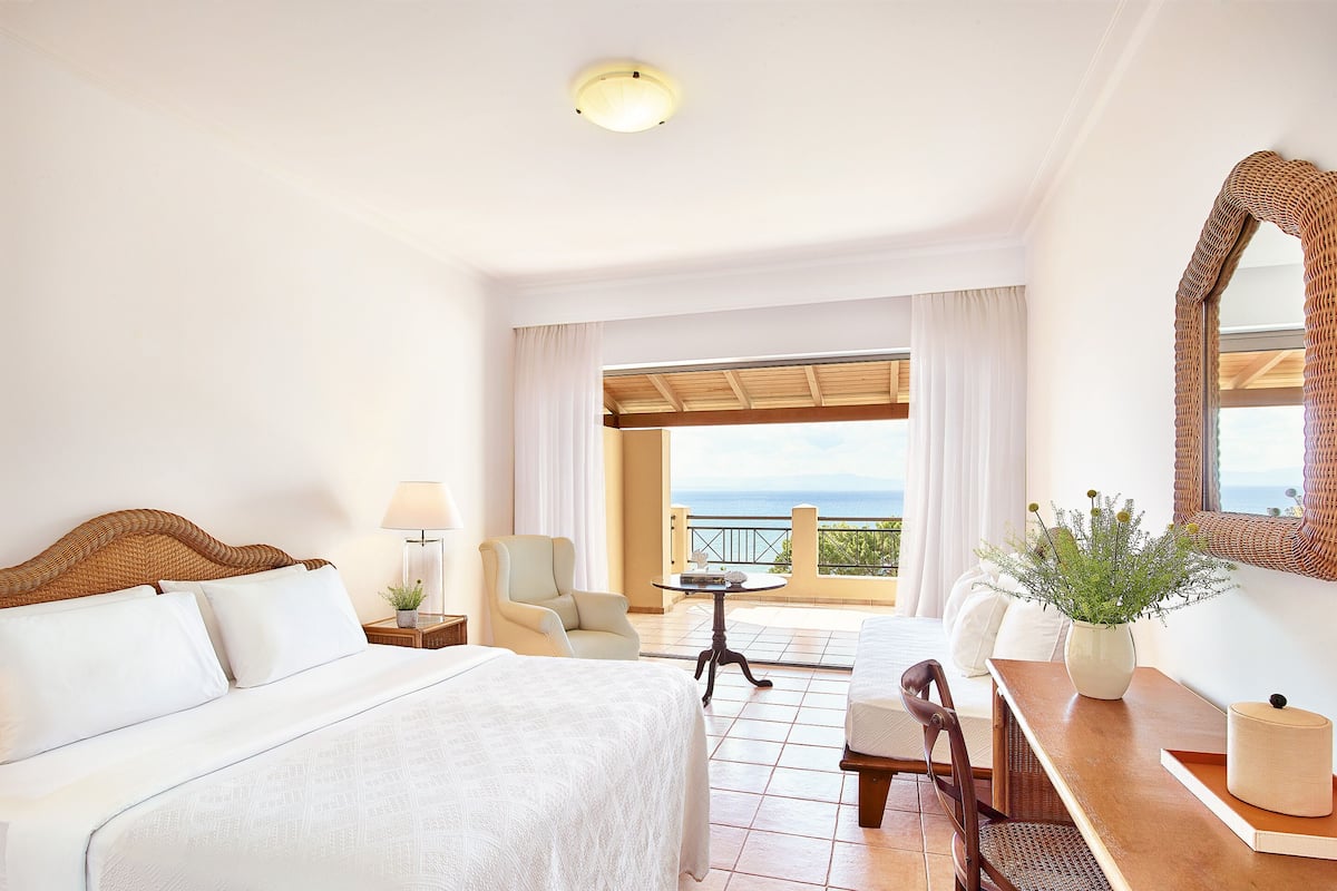 Habitación de hotel con cama, escritorio, silla, mesa, lámpara, planta en maceta y vista al mar.