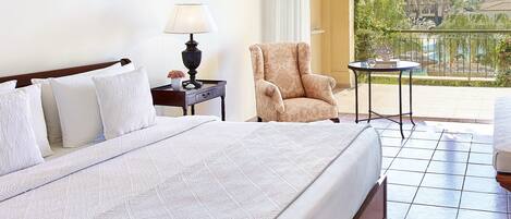 A hotel room with a large bed, a chair, a small table, and a view of a pool and greenery.