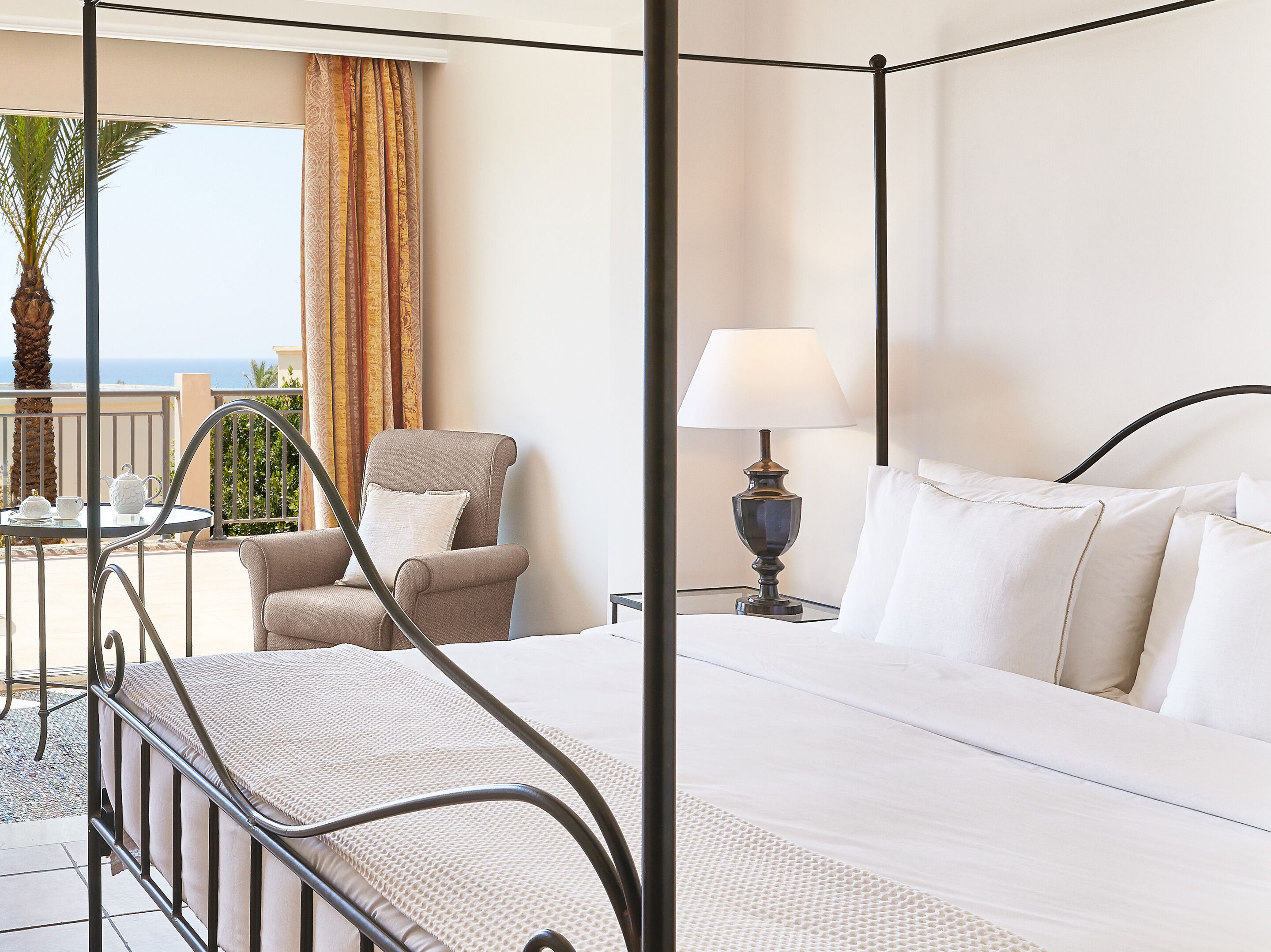 A four-poster bed with a white mattress, a bedside table with a lamp, a chair, and a view of the outdoors.