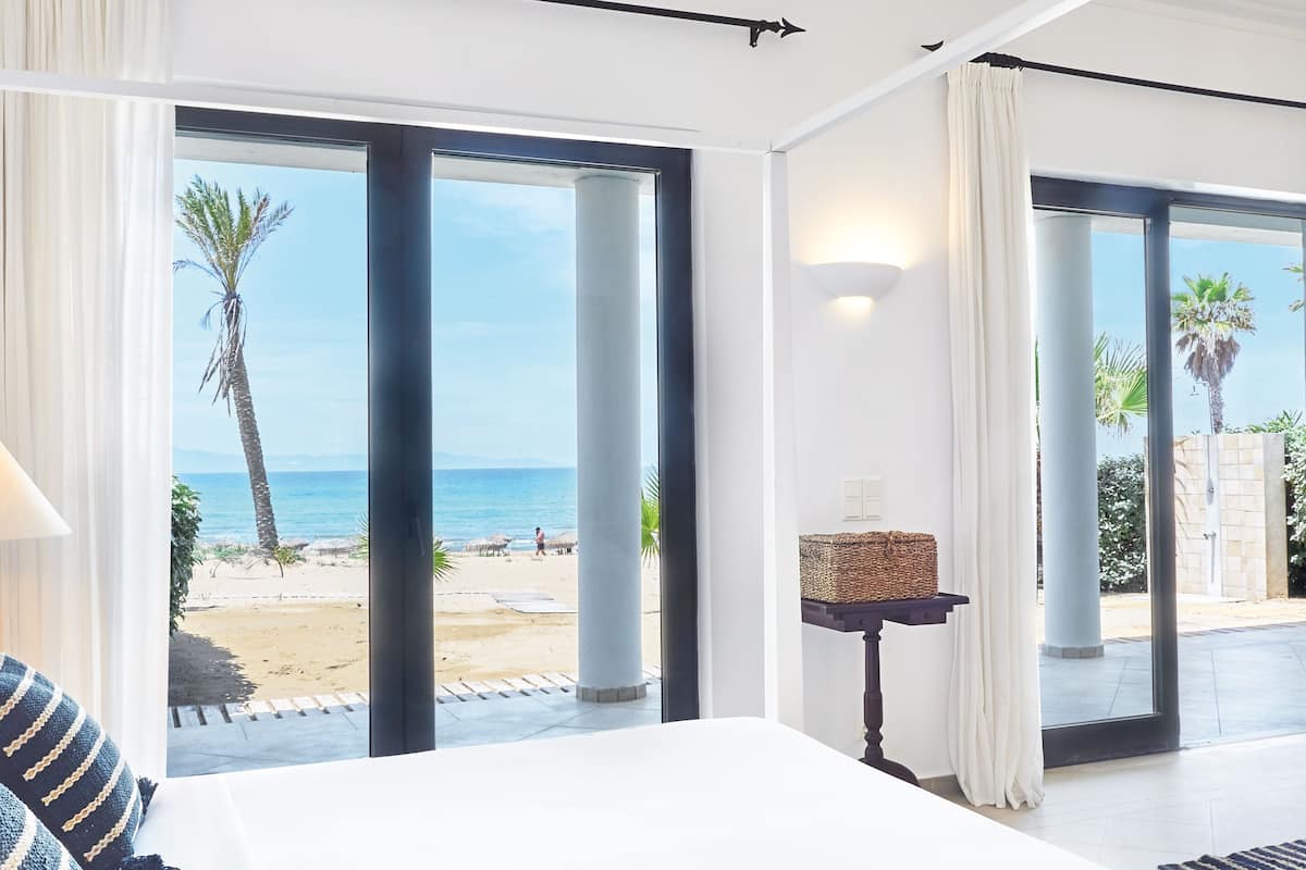 Habitación de hotel con una cama grande, vista a la playa y una pequeña mesa con una lámpara.