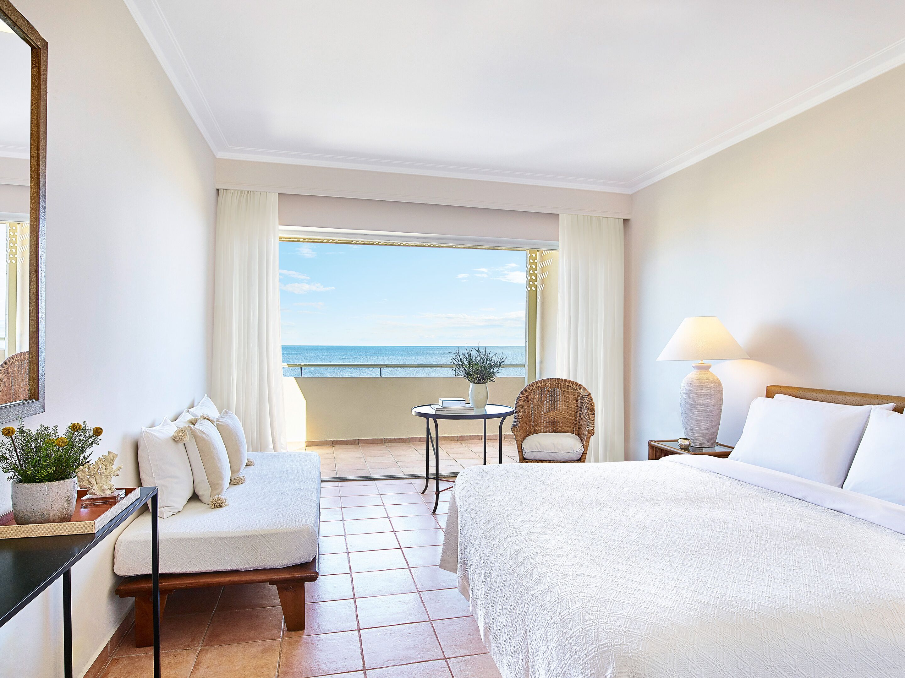 A hotel room with a bed, a chair, a small table, and a view of the sea.